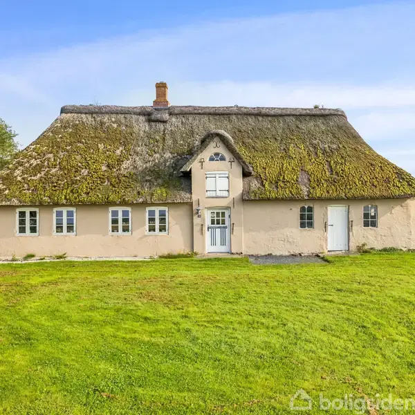 Stråtækt hus står stille i en grønt landskab med græsplæne og træer. Huset har flere vinduer og døre, dets tag er dækket af mos og strå.