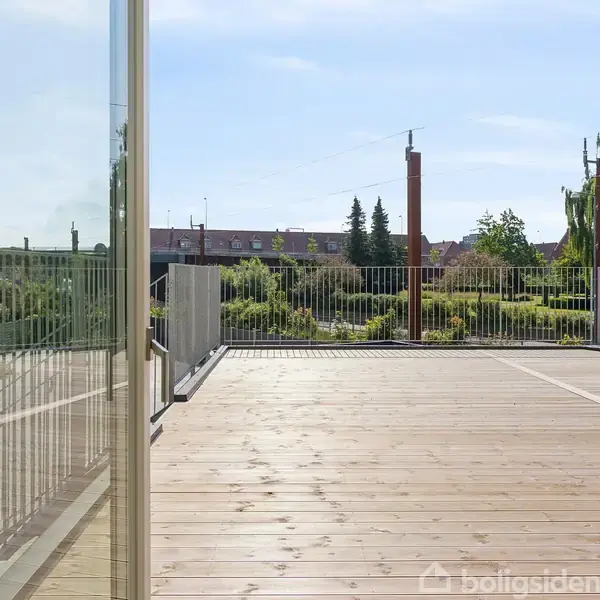 Terrasse reflekterer i stort glasparti, omkranset af moderne metalgelænder. I baggrunden ses grønne træer og et taglandskab, der skaber en urban, men frodig atmosfære.