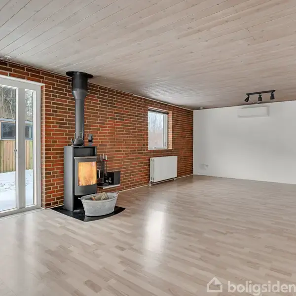 Brændeovn står foran en murstensvæg, opvarmer et rum med lyst trægulv og træloft. Store vinduer til venstre viser sne udenfor.