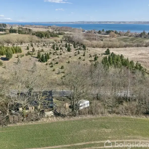 En lille bygning og en campingvogn står blandt træer ved en vej. Baggrunden viser et kuperet landskab med spredte træer og en sø i horisonten under en blå himmel.