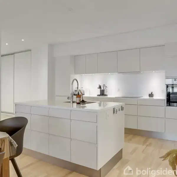 Et moderne køkken med hvide skabe og en stor central ø. Øen har en vask med hane. Rummet er lyst med trægulve, indbygget ovn og spisebord i venstre side.