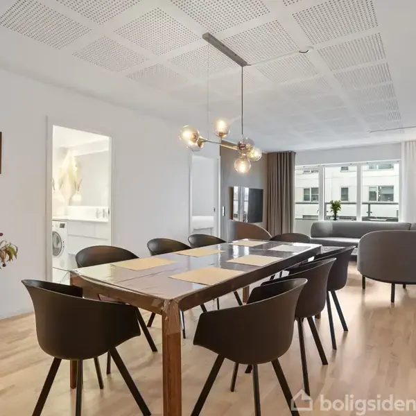 En spisestue med et bord i træ omgivet af otte sorte stole, med en moderne lysekrone hængende over. I baggrunden ses et stueområde med sofaer og et tv.