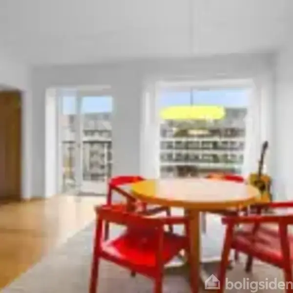 Rundt spisebord med røde stole står i en lys stue med trægulv. Der er en bogreol til venstre og en stor lampe hænger over bordet.