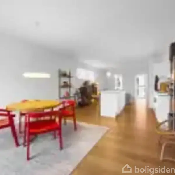 Et lyst rum har et rundt bord med røde stole på et tæppe. Køkkenet ses i baggrunden med bøger på en reol til højre.