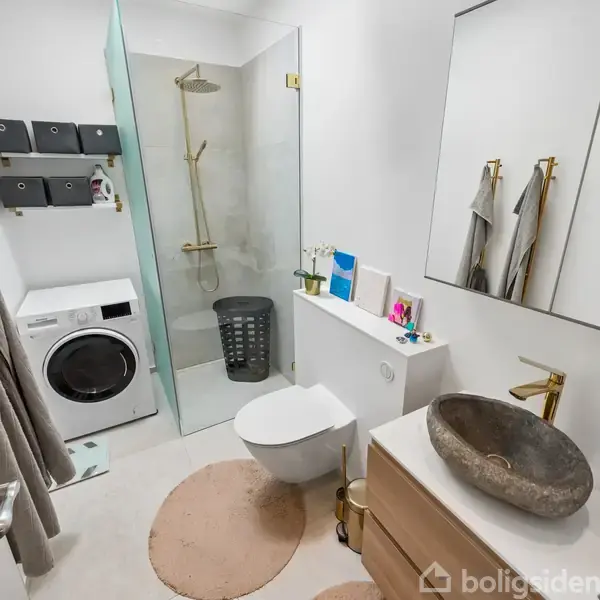 Badeværelse med bruser i glasaflukke, vaskemaskine og beige tæpper. Stenlignende vask med spejl over, håndklædeholder med håndklæder. Hylde med små planter og toiletartikler. Lys og moderne indretning.