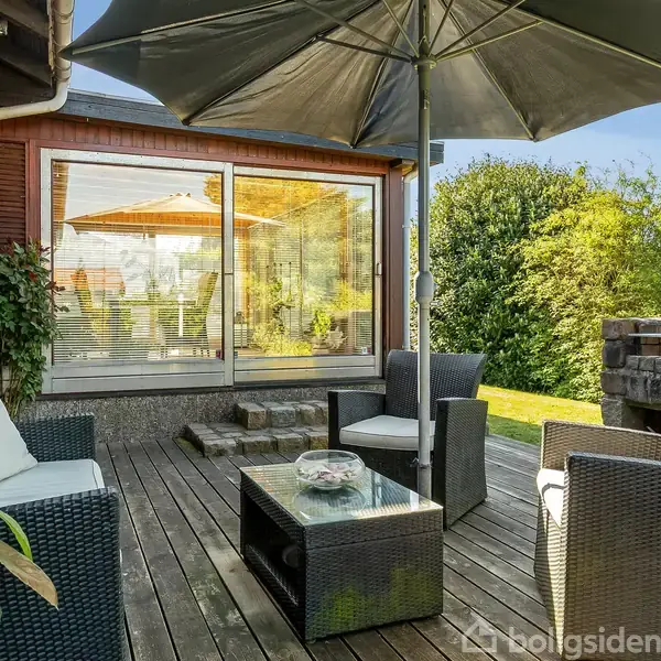 Terrassemøbler står på en træterrasse med en stor parasol. Terrassen er ved et hus med store glasdøre, omgivet af grøn beplantning og stenmur.