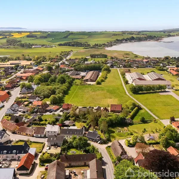 En landsby set fra luften med huse spredt omkring grønne områder og veje. En fjord kan ses i baggrunden med landbrugsmarker omkring.