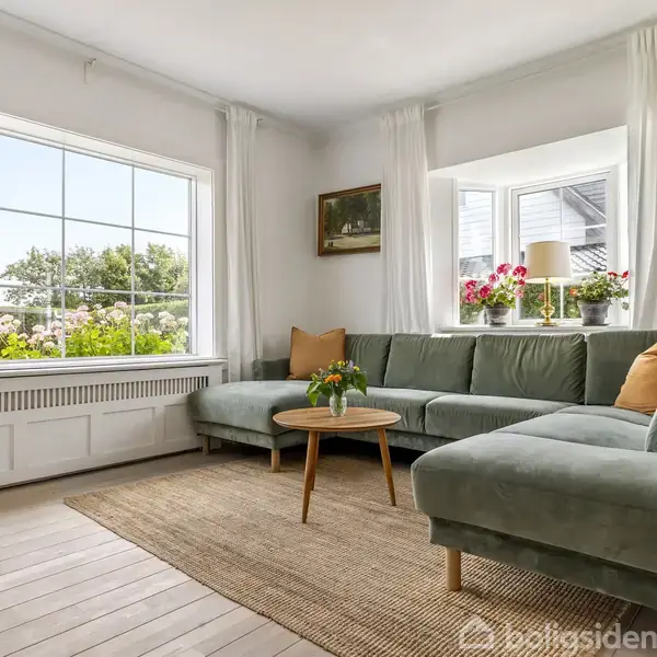 Grønt sofasæt står foran et stort vindue, udsigt til have. Stuebord med blomster på tæppe, trappe til højre. Rummet er lyst med hvide vægge og gardiner.