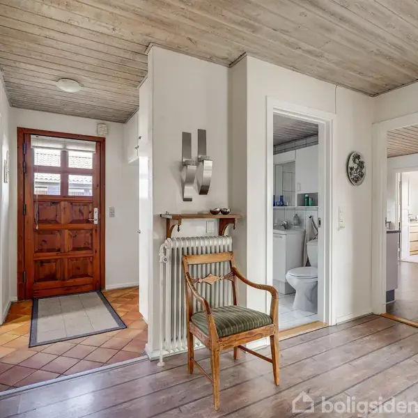 En entré med en træstol foran en radiator. Der er en åben dør til et badeværelse og indgang til et køkkenområde. En rødbrun hoveddør ses i baggrunden.