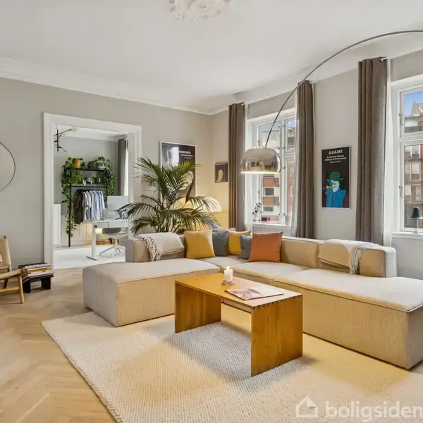 Stor, lys stue med beige hjørnesofa omkring et træbord. En buet gulvlampe hænger over sofaen. Store vinduer med udsigt til lejligheder. Plakater og planter skaber en moderne atmosfære.
