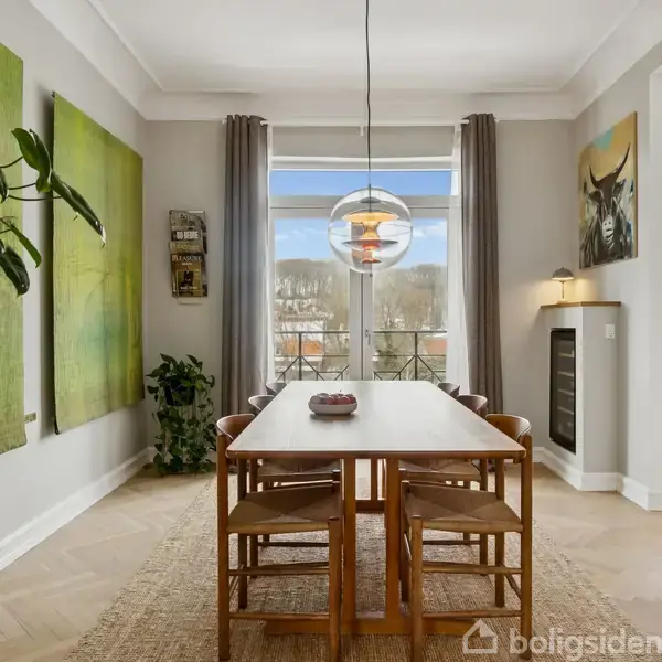 Spisebord med seks stole står i et elegant, lyst rum med store vinduer og grønne kunstmalerier på væggen. En moderne lampe hænger over bordet, og planter dekorerer hjørnerne.