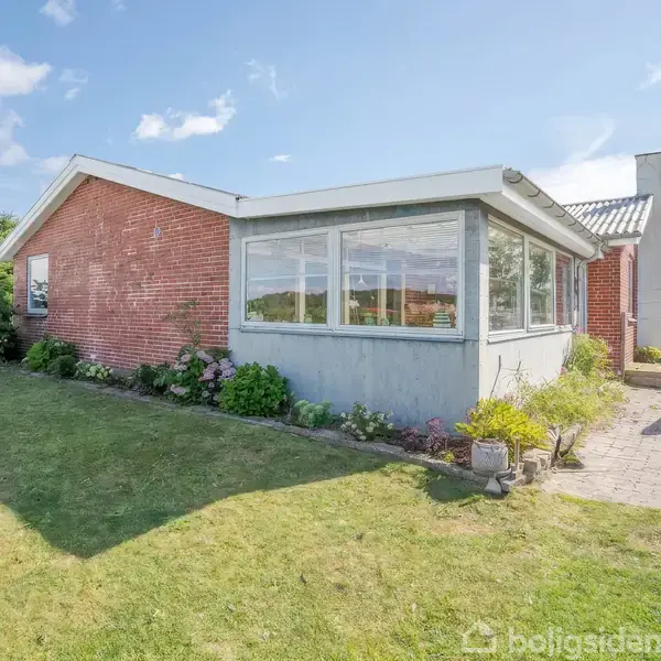 Rødt murstenshus med stor glasveranda, omgivet af grøn græsplæne og enkelte buske, i et solrigt boligområde med en terrasse til højre med havemøbler.