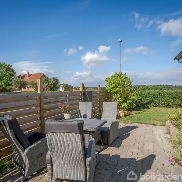 Bord med seks kurvestole står på en terrasse af fliser. Lige ved siden er en træhegn. Grønne planter og græsplæne findes i baggrunden under en blå himmel.