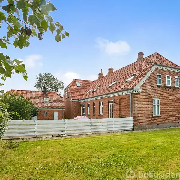 Et rødt murstenshus med skråt tag står stille i en frodig have med græsplæne. Det er omgivet af et hvidt hegn, og træer ses i baggrunden under en blå himmel.