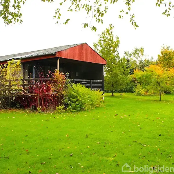 Et rødt og sort træhus står stille i en frodig have med grønne træer og buske omkring. Græsplænen er tæt og velplejet.