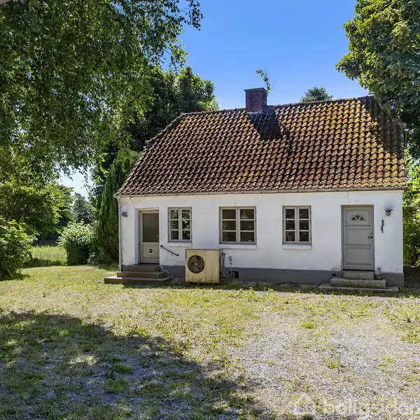 Et lille hus med et rødt tegltag står i et grønt, skovomgivet område. To døre og fire vinduer pryder facaden. Grusvej foran huset er omgivet af træer og buske.