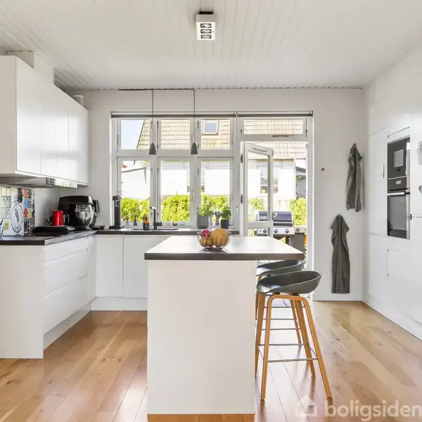 Køkken med hvidt interiør og sort bordplade, arrangeret omkring en central ø med barstole. Store vinduer giver udsigt til terrasse og grønt hegn udenfor.