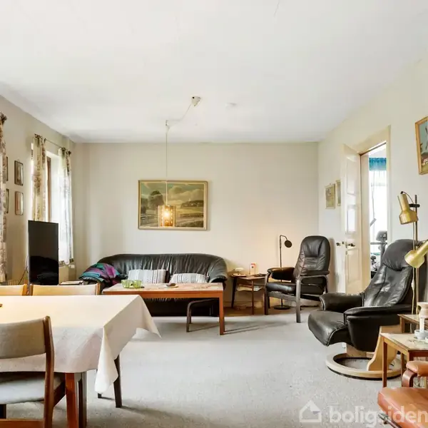 En stue med sort lædersofa og lænestol, placeret nær et rundt sofabord. Ved siden af står et spisebord med stole. Rummet har flere vinduer og vægkunst.