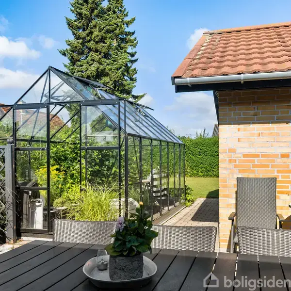Et drivhus står i en have ved siden af et murstenshus. Foran er der havemøbler og planter på et terrassebord. Himlen er blå med spredte skyer.