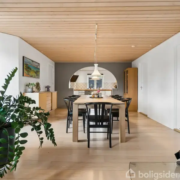 Spisebord omgivet af sorte stole står i en spisestue med lyst trægulv og loft. En stor plante og et brændekomfur ses i rummet, samt kunst på væggene.