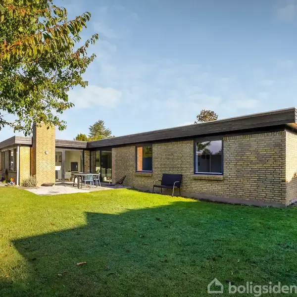 En gul murstensvilla står på en græsplæne med træer omkring. En terrasse foran huset har et bord og stole, og solen skinner på murene og græsset.