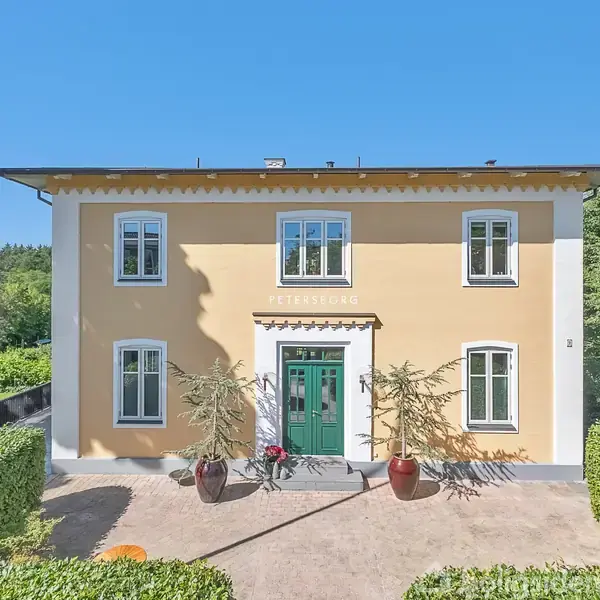 En gul villa med navnet "PETERSBORG" over grønt hoveddør. Døre flankeres af planter i store krukker. Omkranset af hække og træer, ligger under en klar blå himmel.