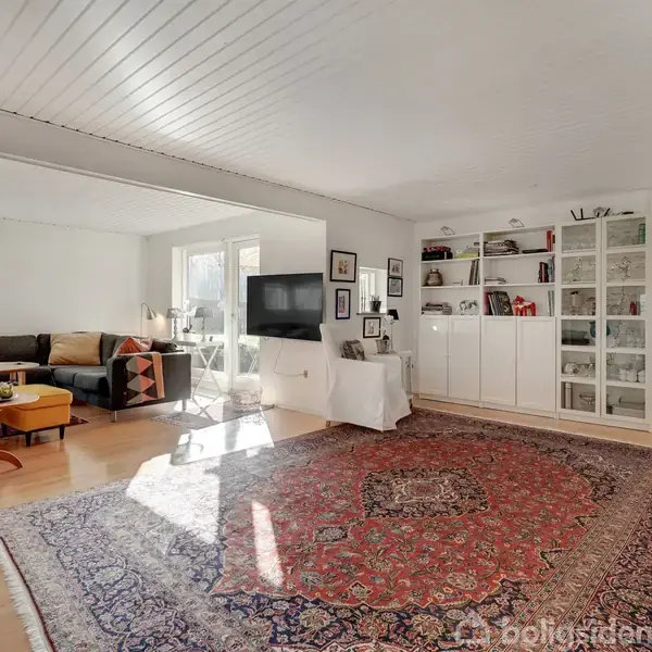 Stuen har en stor orientalsk tæppe, en hjørnesofa med farverige puder, og en gul lænestol med fodskammel. En reol med glaslåger står ved væggen, mens et TV hænger tæt på vinduet.