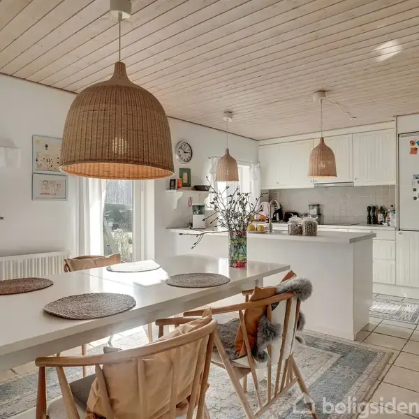 Spisebord med stole har dækkeservietter og blomster. Over dem hænger kurvelamper. Køkken med hvide skabe og apparater er i baggrunden. Gulvtæppe og fliser i et lyst rum.