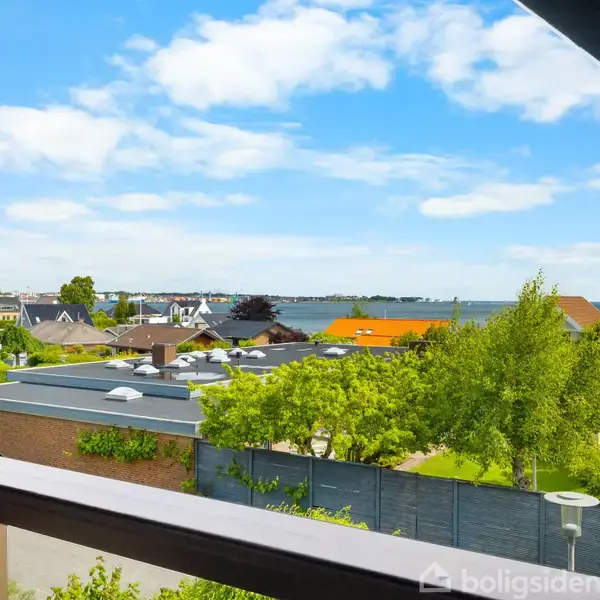 Udsigt fra balkon over en kystby med huse omgivet af grønne træer. Havet ses i baggrunden under en himmel med spredte skyer. Ingen tekst er til stede.