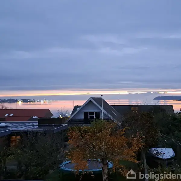 Forskellige hustage og træer danner forgrund for en udsigt over en rolig vandflade ved skumringstid. Lyset reflekteres delvist på vandet, og himlen er overskyet.