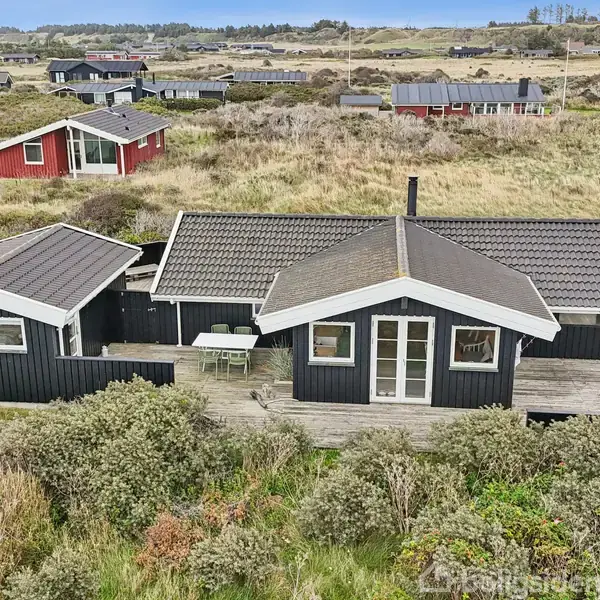 Sort sommerhus med gråt tag står stille omgivet af klitter og andre huse i baggrunden. Terrasse med bord. Nordlig kystmiljø med lav vegetation.