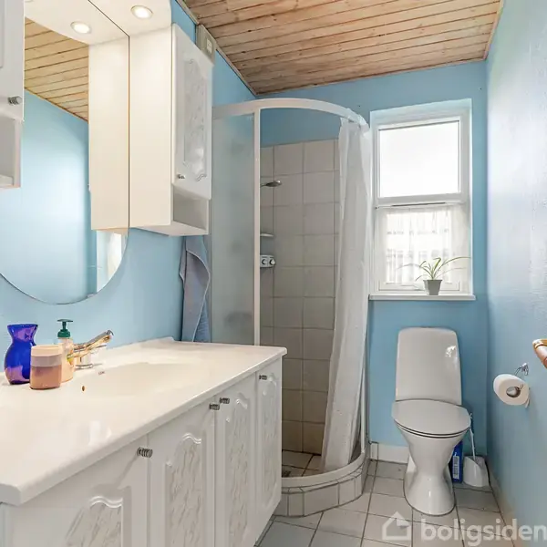Badeværelse med blå vægge, hvide møbler og indrettet med et rundt spejl. Brusekabine i hjørnet, toilet ved vindue og håndvask med sæbe og glas på bordpladen.