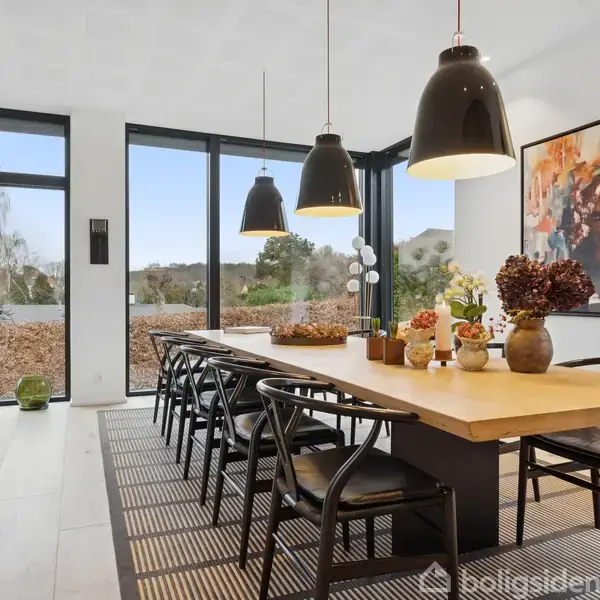 En stor træspisebord med sorte stole står i et lyst spisestue. Bordet er dekoreret med planter og kunstgenstande. Store vinduer med udsigt over en have omgiver rummet.