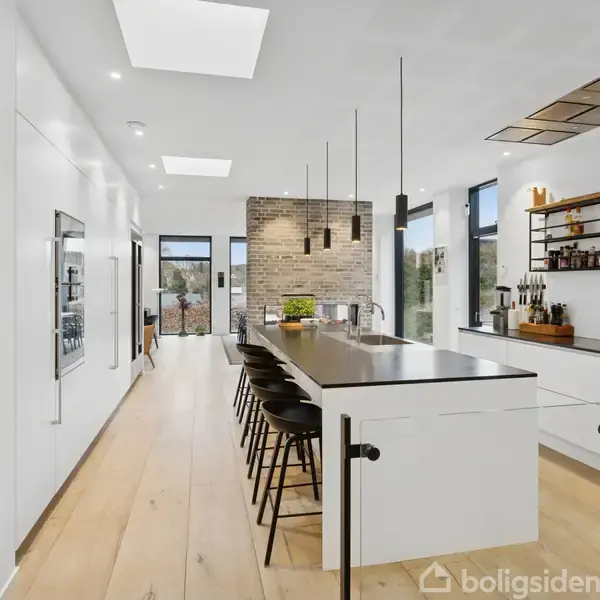 Et moderne køkken med en lang spiseø fyldt med sorte stole og en stor hvid bordplade. Omgivet af hvide vægge, kunst på væggen, og hylder med dekoration.