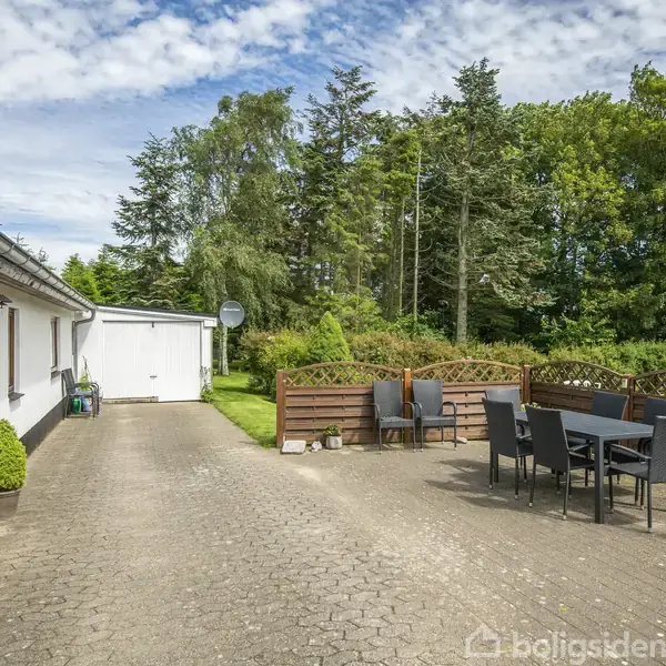 Hvide husfacade med stenbelagt gårdsplads og gruppe havemøbler ved siden af træhegn. Nabogarage og frodigt skovområde i baggrunden under en delvist skyet blå himmel.