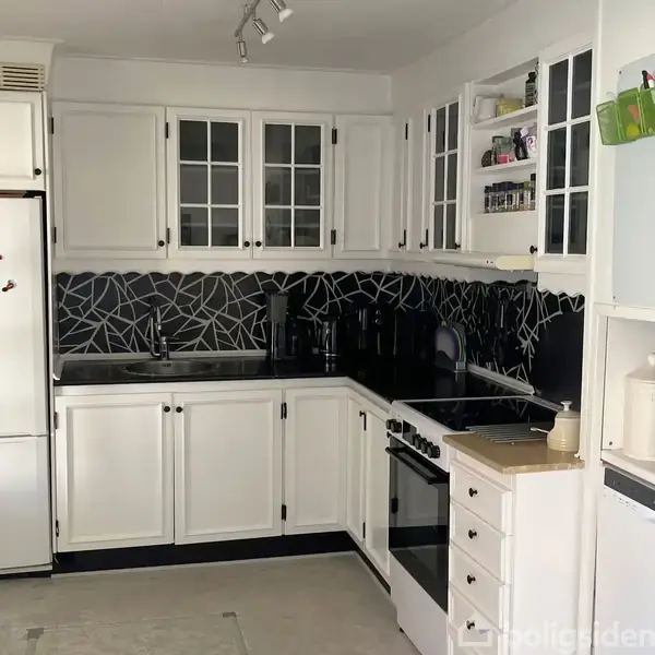 Et hvidt køkken med sorte detaljer har en komfur, ovn og opvaskemaskine. Køleskabet står til venstre. Bagvæggen har et sort mønster, mens en mikrobølgeovn sidder til højre.