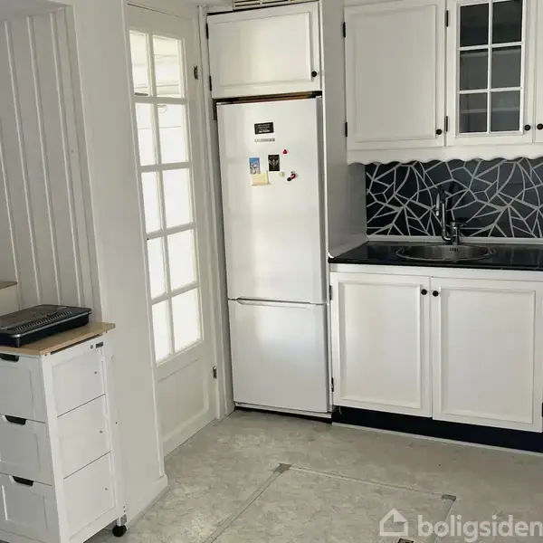 Køleskab står ved siden af hvide køkkenskabe, som har en sort mosaik-flisevæg. En fortrolig printer er placeret på en rullevogn til venstre. En glasdør findes i baggrunden.