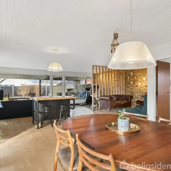 Et rundt træbord med stole står i forgrunden. I baggrunden ses en stue med store vinduer, sofaer og en barafdeling med høje stole. Plante og lamper som dekoration.