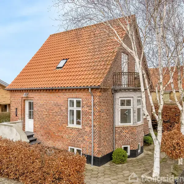 Et rødstenshus med rødt tegltag står stille ved siden af en fortov. Huset har en lille altan og er omgivet af en hæk og et fortov.