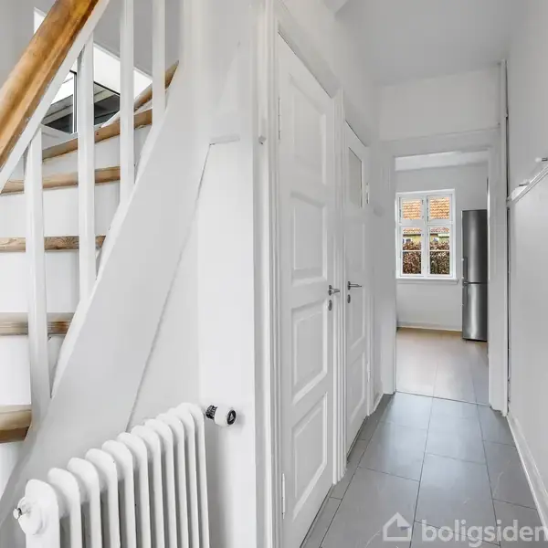 Trappe med trærækværk fører opad; gangen er hvide vægge og gråt gulv; en radiator på venstre side og flere døre fører til andre rum.