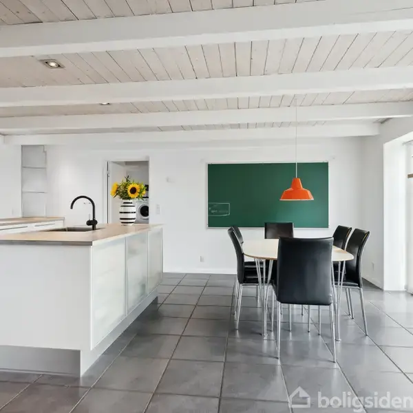 Køkken-alrum med hvid køkkenø og sort armatur. En spiseplads med sort bord og stole under orange lampe. Solsikker står på køkkenbordet, og der er store vinduer omkring.