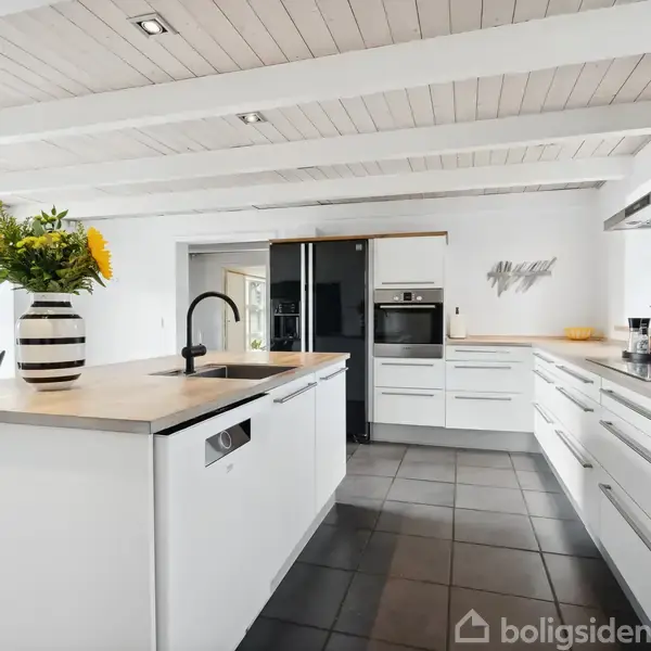 Et køkken med en hvid ø med sort stribe vase med solsikker; moderne ovn og køleskab i baggrunden. Store vinduer giver udsigt til haven.