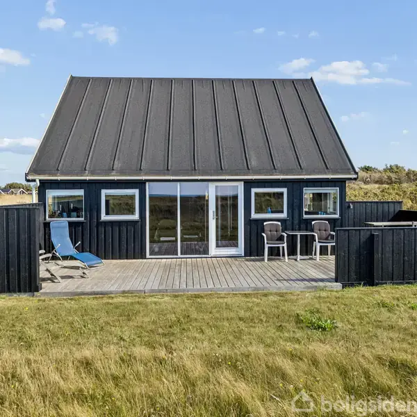 Et sort sommerhus med store vinduer og skråt tag står på en træterrasse. To stole og en solseng er placeret på terrassen. Huset er omgivet af grønne marker og lys himmel.