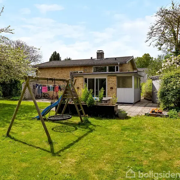 Et gyngestativ med en redegynge står i en grøn have foran et hus med terrasse og store vinduer. Træer omgiver området, og haven ser velplejet ud.