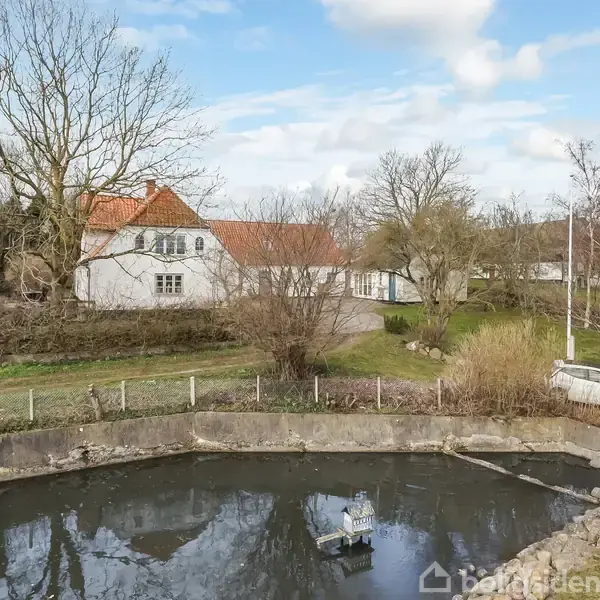 En hvid bygning med rødt tag står ved siden af en lille, indhegnet dam. Træer omgiver det naturskønne område, og en grusvej løber forbi. En parkeret bil ses nær dammen.
