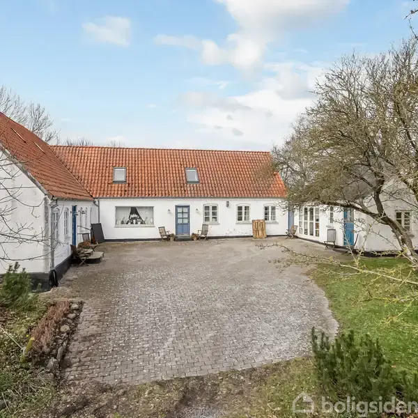 Hvidt, L-formet hus med rødt tegltag står omkring en brolagt gårdsplads, omgivet af spredte træer og græs. Skyfri blå himmel baggrund. Ingen tekst er til stede.