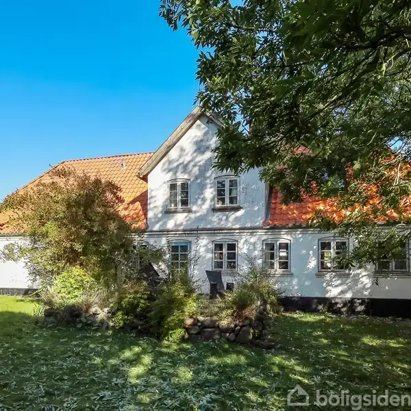 Et hvidt, muret hus med rødt tegltag og flere små vinduer står fredfyldt i en frodig have med grønne træer og græs, badet i solskin under en klar blå himmel.