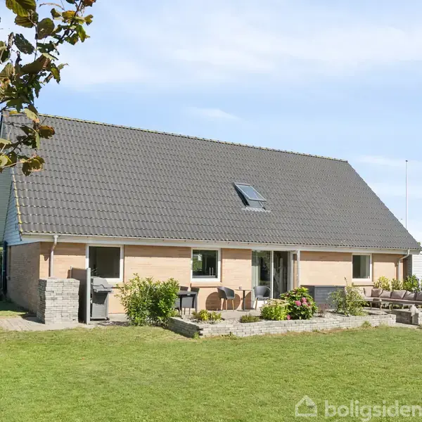 Et murstenshus står stille i en velplejet have med græsplæne. Huset har et skråt tag med en enkelt kvist og store vinduer, omgivet af buske og træer.