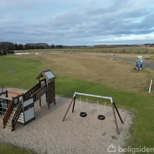 Legeplads med et klatretårn og to gynger placeret på sandbund. Aktion: Friluftslegeplads. Kontekst: Omgivet af græs, asfaltvej og åbent landskab under en overskyet himmel.