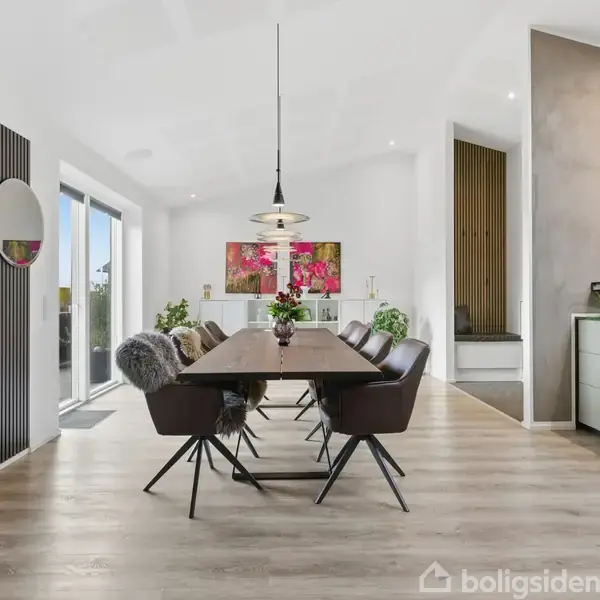 Et moderne spisestue med et langt træbord omringet af seks stole. Til venstre brænder en pejs. Rummet er lyst med store vinduer, planter og kunstværker på væggene.